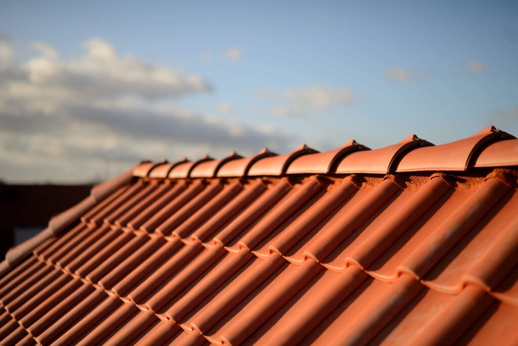 Close-up of a terracotta tile roof under a clear blue sky with scattered clouds. The angled sunlight emphasizes the texture and neat alignment of the tiles, much to a roofer's delight, creating a warm, serene atmosphere reminiscent of contrasting flat roofs in urban settings.