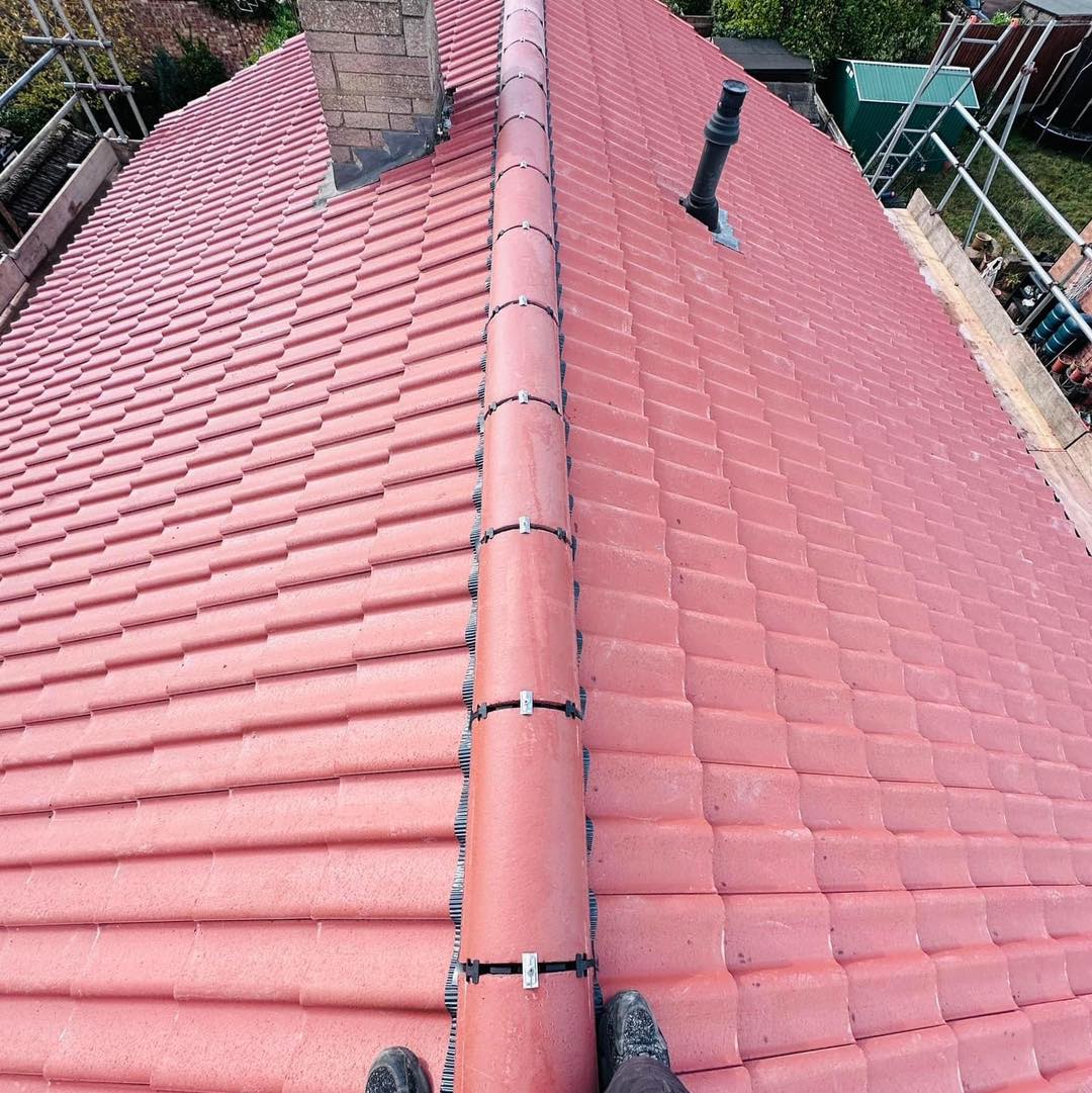 The view features a roof with red tiles, a chimney, and a vent pipe. A roofer's boots appear at the bottom, indicating work on new roofs or possibly replacement guttering. Scaffolding and greenery enhance the scene in the background.