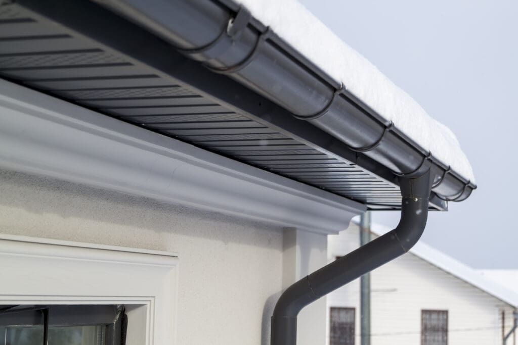 Close-up of a house roof with gray replacement guttering, partially covered in snow. A drainpipe extends downward, complementing the light-colored wall and window frame beneath. The overcast sky adds a gloomy touch, hinting at the roofer’s craftsmanship with flat roofs in challenging weather.