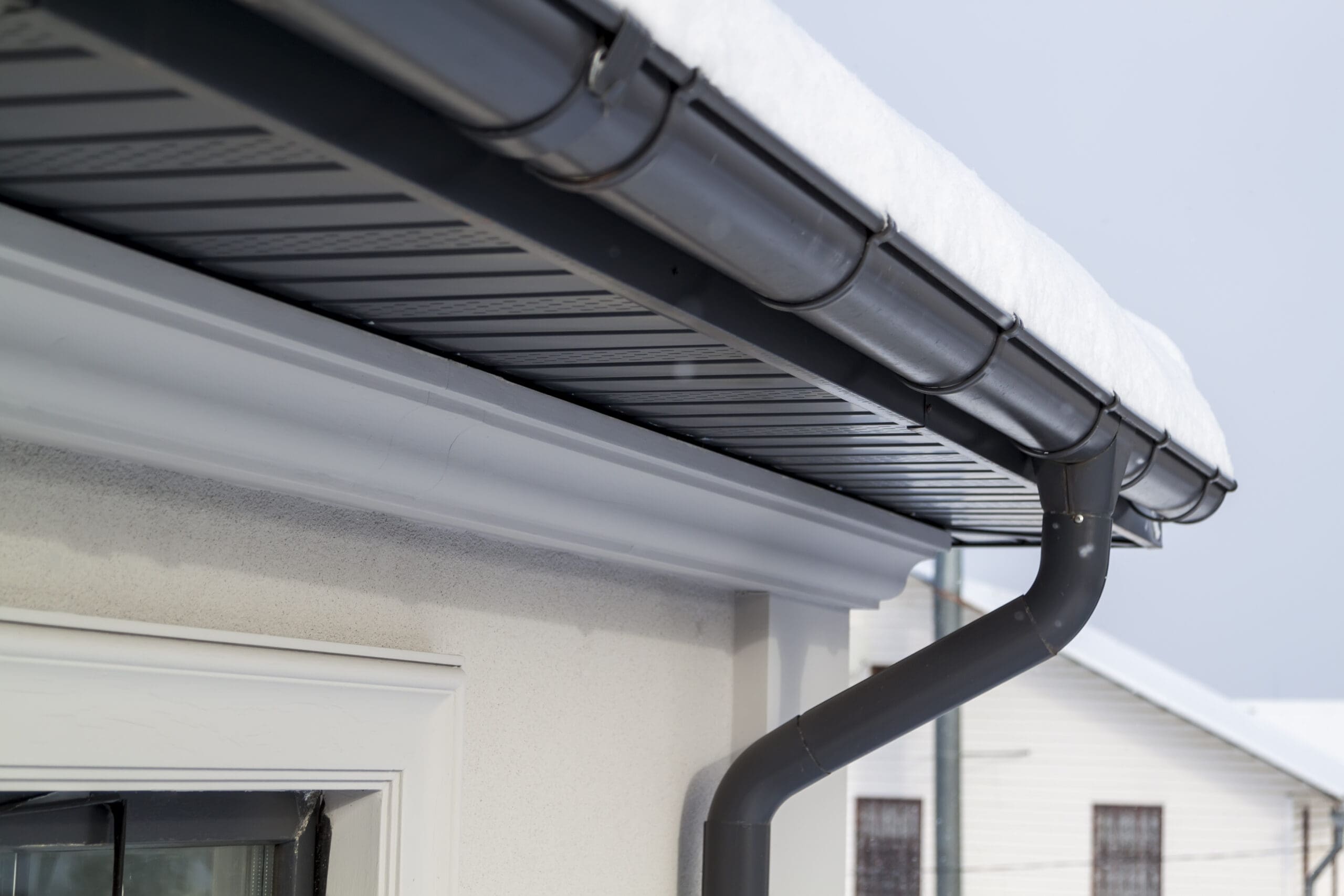Close-up of a house roof with gray replacement guttering, partially covered in snow. A drainpipe extends downward, complementing the light-colored wall and window frame beneath. The overcast sky adds a gloomy touch, hinting at the roofer’s craftsmanship with flat roofs in challenging weather.
