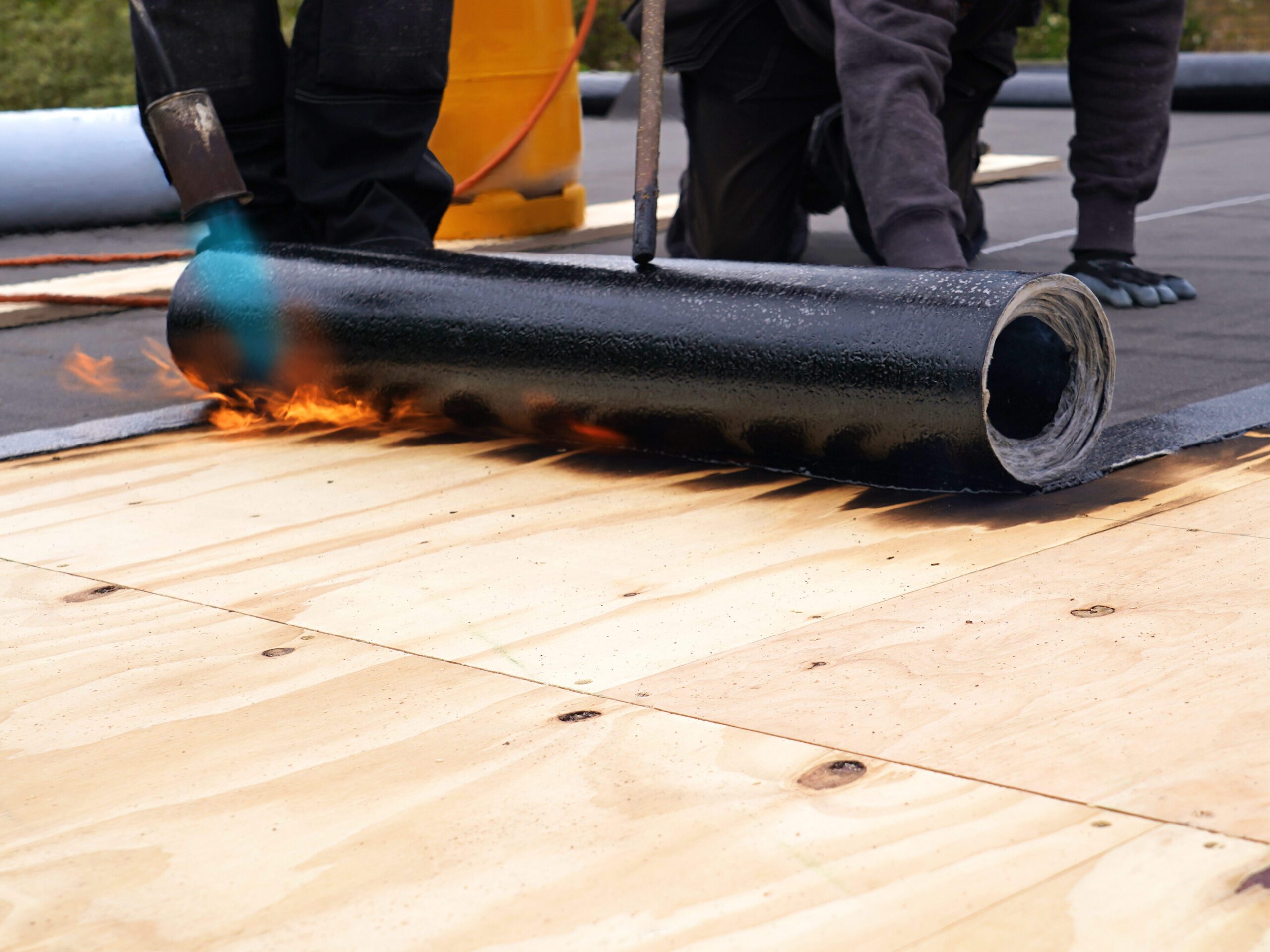 Two roofers apply a roll of tar paper onto a plywood flat roof using a propane torch. One holds the roll while the other uses the torch, creating a small visible flame. In the background, part of a gas cylinder is visible.