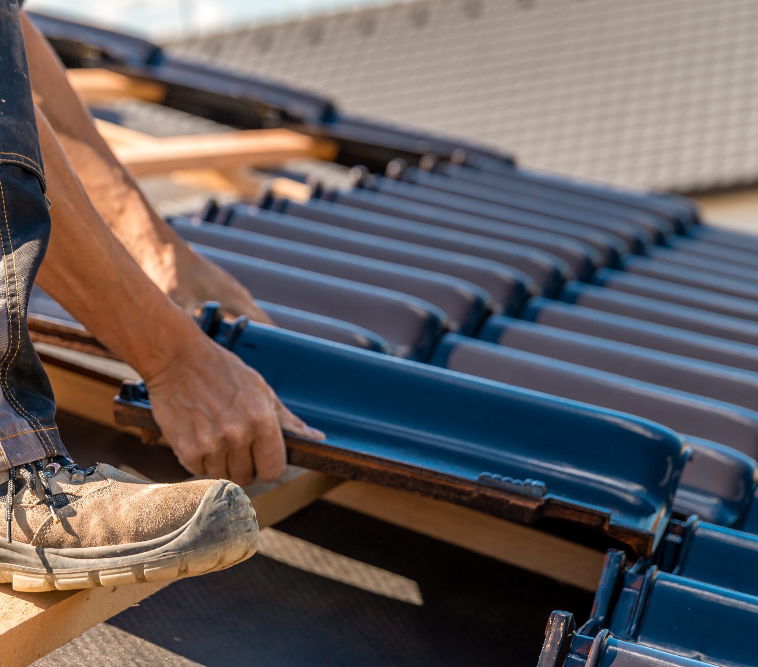 A skilled roofer's hands and feet align blue roofing tiles on a sloped wooden framework. The worker, wearing tan safety shoes, moves confidently as the sun casts shadows on the roof surface. The partially tiled roof stretches diagonally across the image.