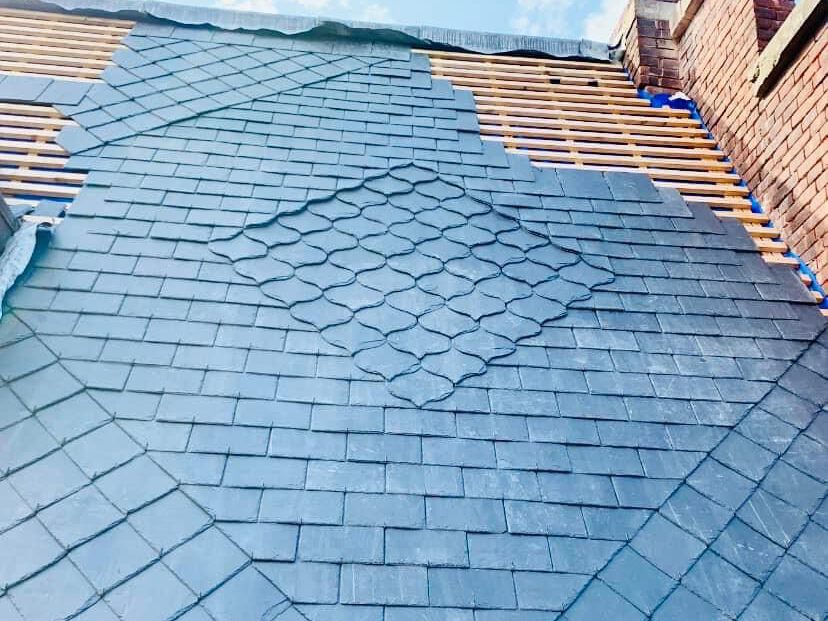 A skilled roofer is crafting a stunning new roof with slate tiles arranged in interlocking diamond shapes. The grayish-blue pattern adds elegance, while the exposed wooden battens at the top hint at ongoing progress.