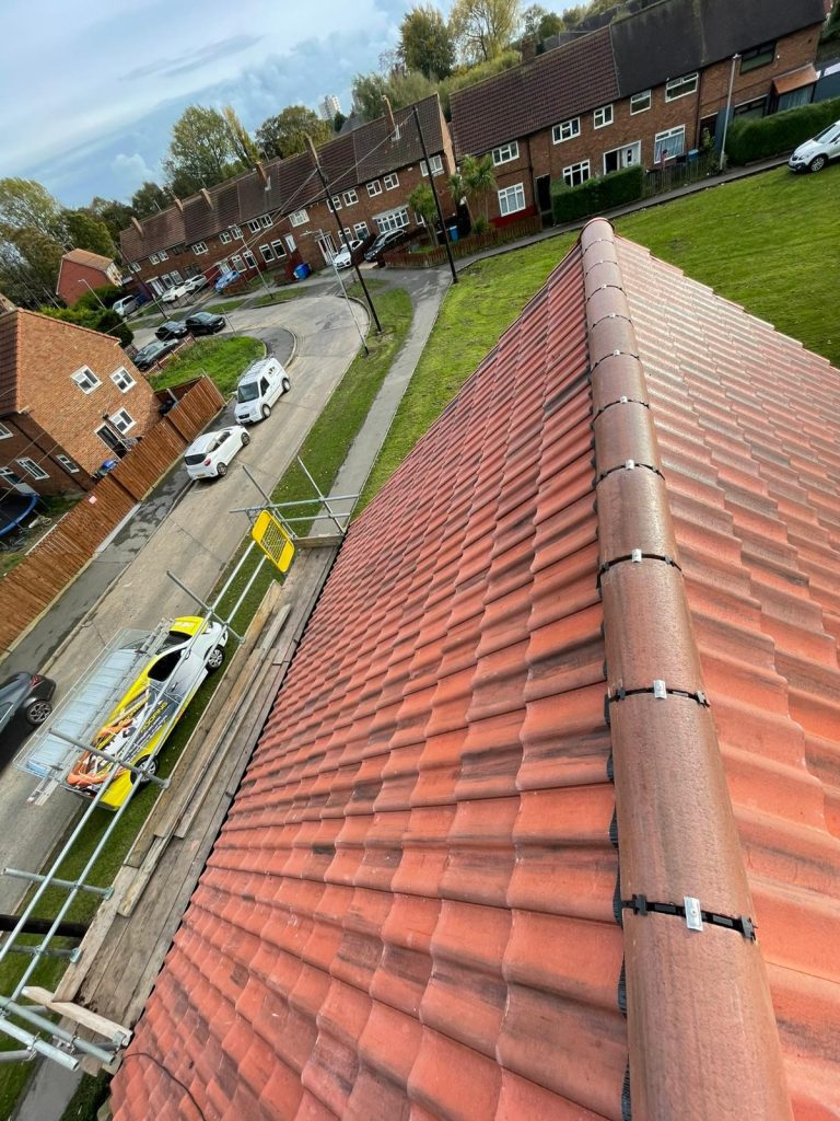 A view from a rooftop with reddish tiles, overlooking a suburban street with brick houses, parked cars, a grassy area, and scaffolding beside the building. The sky is partly cloudy.