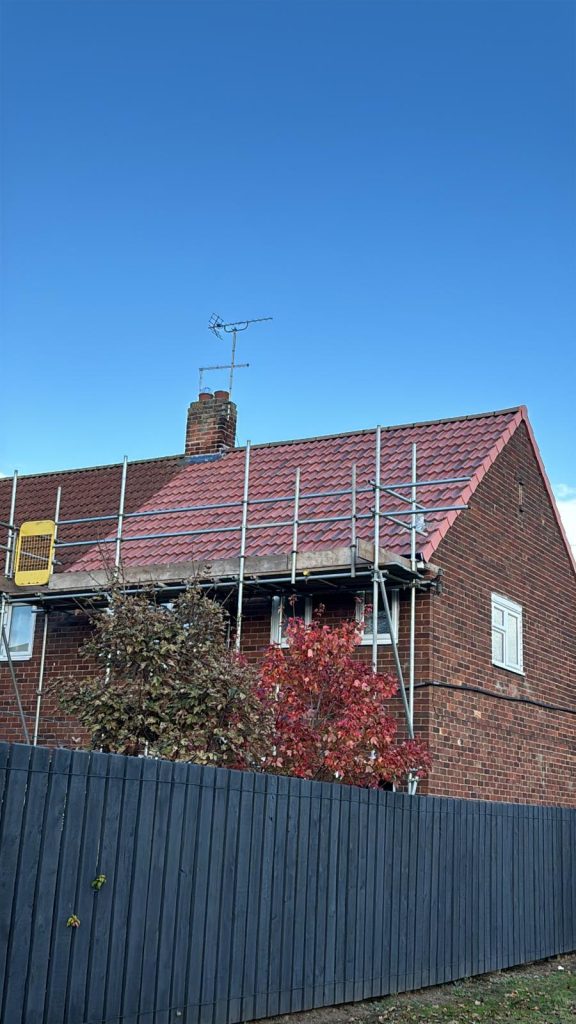 A brick house with red roof tiles, scaffolding along one side, and a TV antenna on the chimney. A tall dark fence and a tree with red leaves are in front, under a clear blue sky.