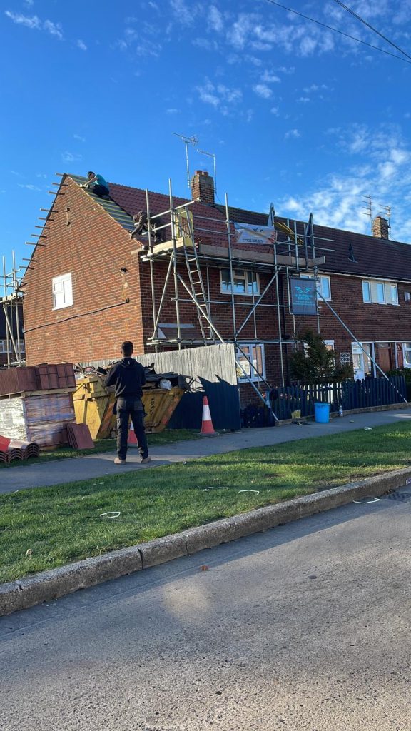 A worker stands on the ground near a brick house under construction, while another person works on scaffolding set up against the roof. A blue sky with clouds is visible above. Construction materials are stacked nearby.