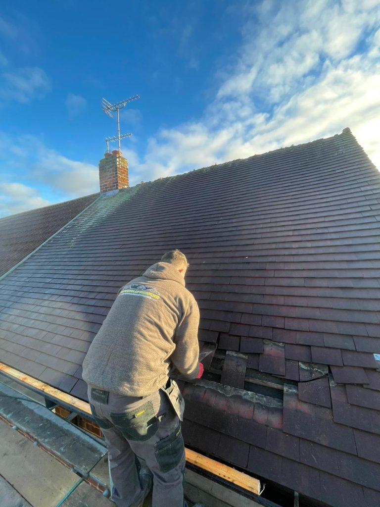 A person wearing a gray hoodie is repairing dark brown roof tiles on a house under a blue sky with scattered clouds. An antenna and a brick chimney are visible in the background.