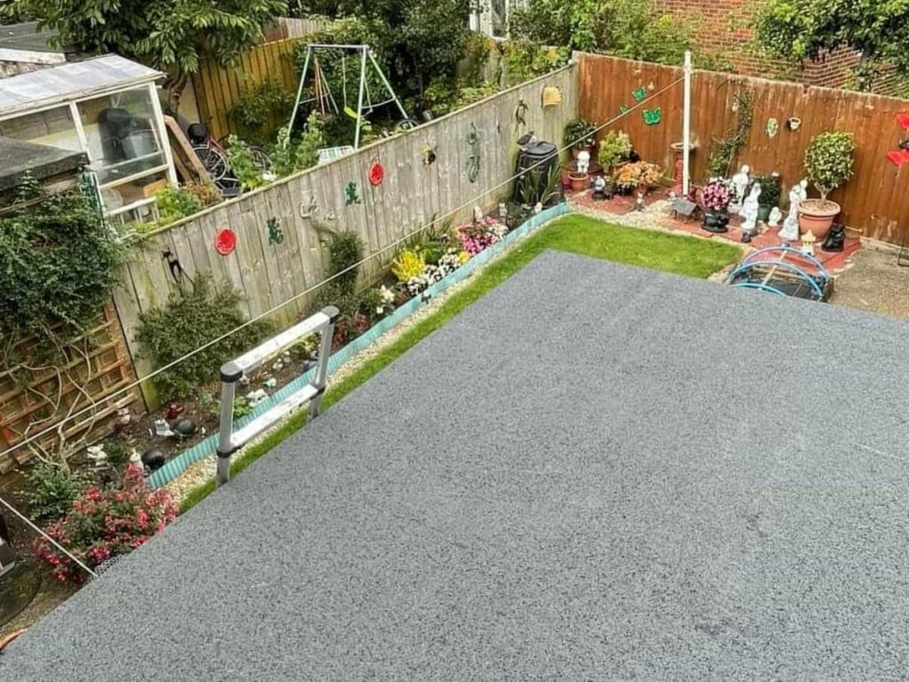 A view of a colorful backyard garden with plants, flowers, lawn ornaments, statues, a swing set, and a small grassy area bordered by a gray flat rooftop in the foreground.