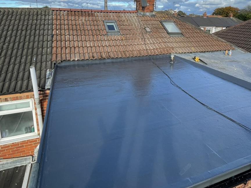 A view of a house roof with a section covered in smooth, shiny dark material next to traditional red and brown tiled roofs. There are two skylights on the tiled roof and a window on the left wall below.