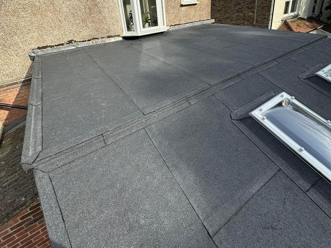 A flat roof covered with dark gray roofing material, featuring two skylight windows and bordered by brick and stucco walls of adjacent buildings.