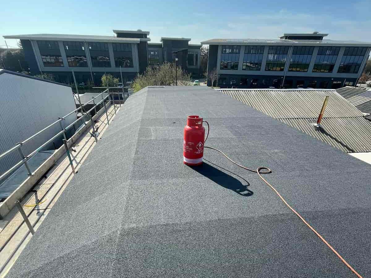 A flat, newly waterproofed roof with a red pressure sprayer sits in the center. Buildings and trees are visible in the background under a clear blue sky.