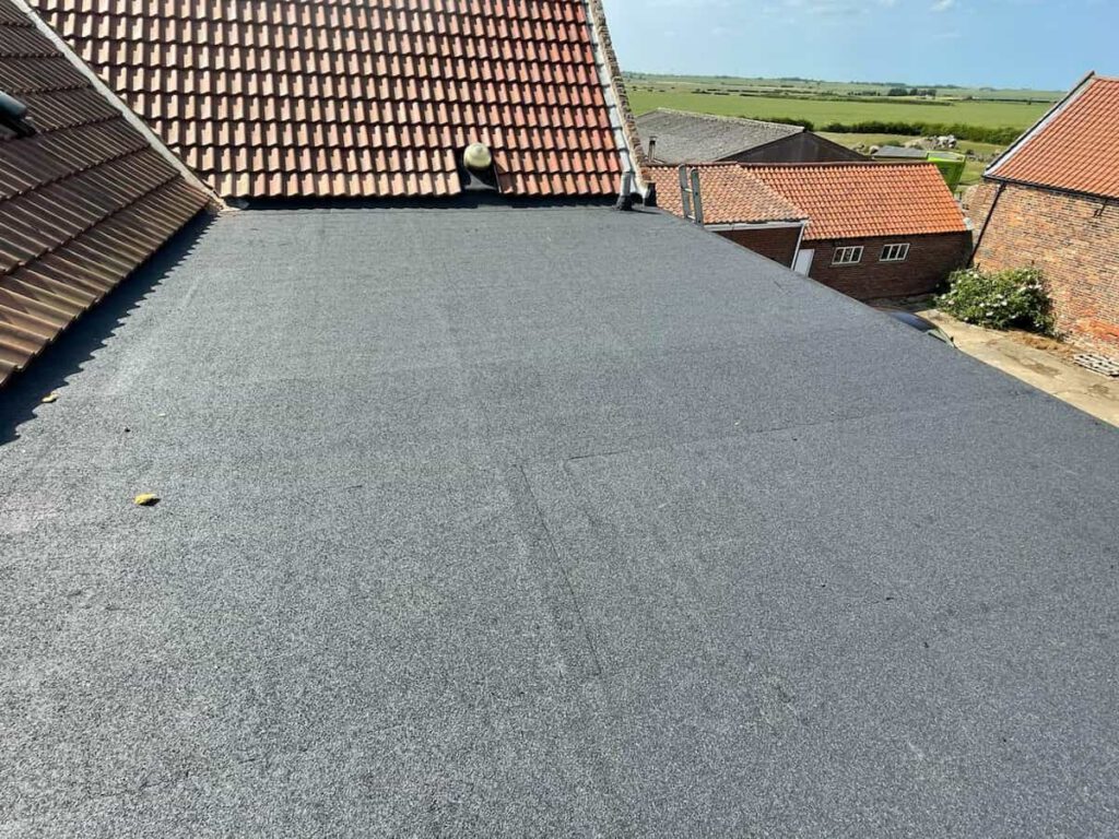 A flat, newly installed black asphalt roof sits between sloped red-tiled roofs on a building overlooking a rural area with fields and scattered brick buildings under a blue sky.