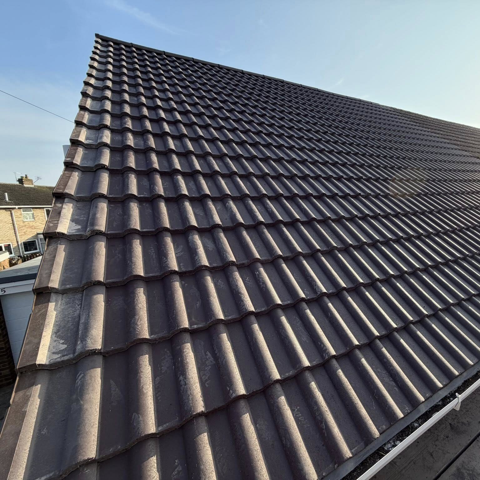 Close-up view of a sloped roof covered with brown, wavy roof tiles under a clear blue sky, with parts of neighboring houses visible at the edge of the image.