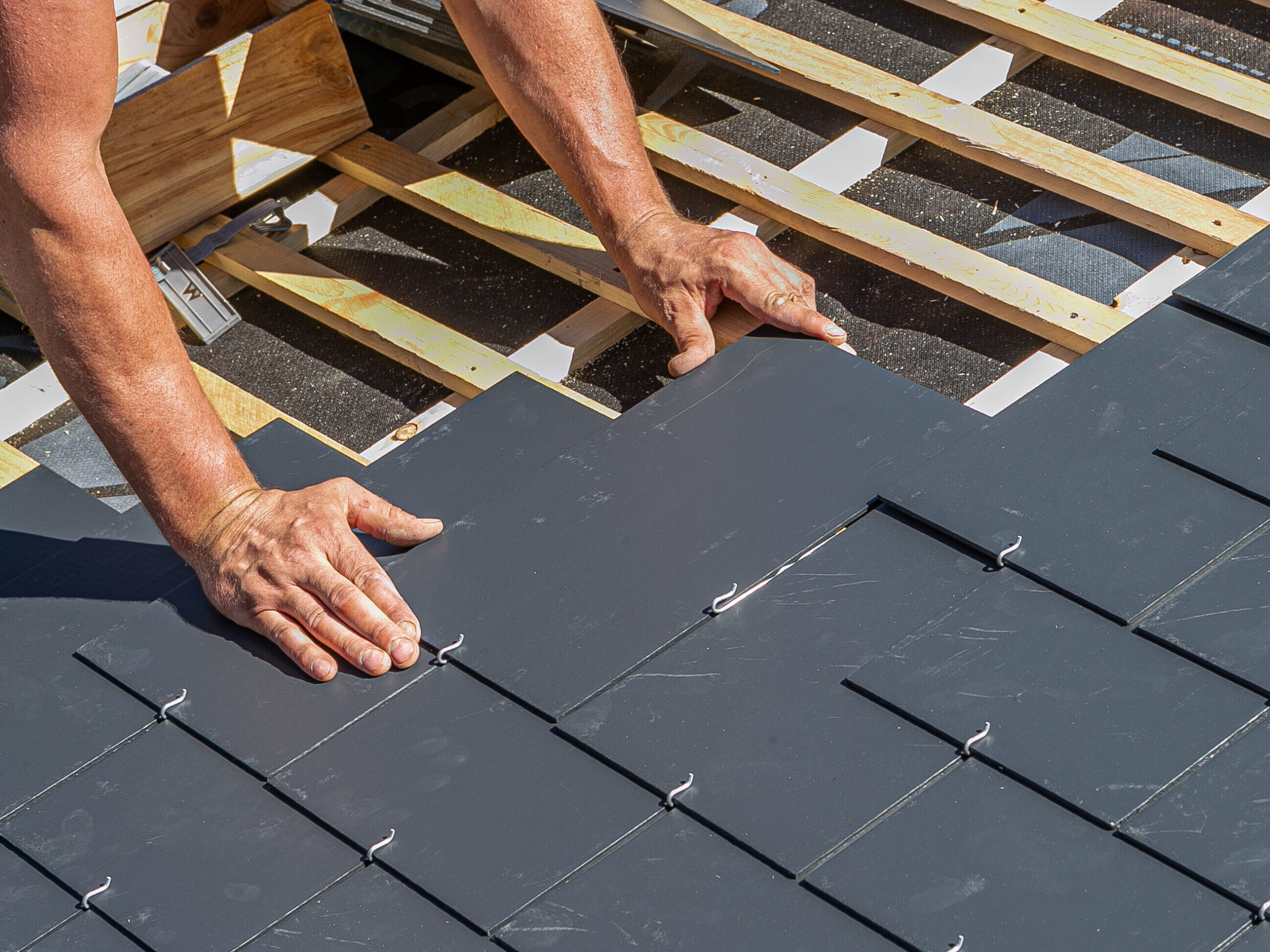 A person in a red shirt is installing dark slate tiles on a new roof. Wooden battens peek from under the incomplete section as they adjust a tile with both hands near its edges. Sunlight casts shadows, hinting at the precision required for this task.