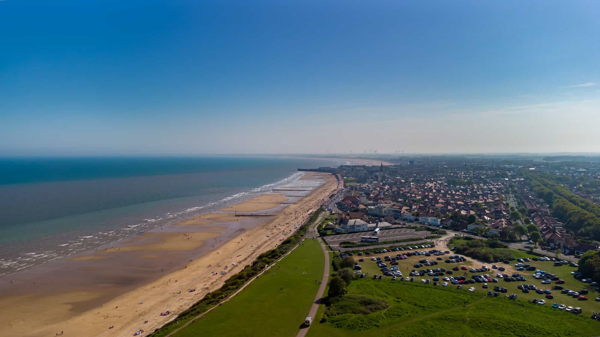 Aerial view of a sandy beach with gentle waves, a coastal town, parked cars, and green fields under a clear blue sky.