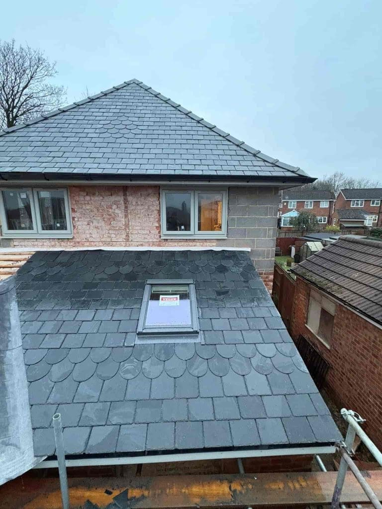 A partially constructed house with a sloped, dark grey slate roof, a window in the upper floor wall, and a roof window marked with a sign. Surrounding the house are other homes and overcast skies.