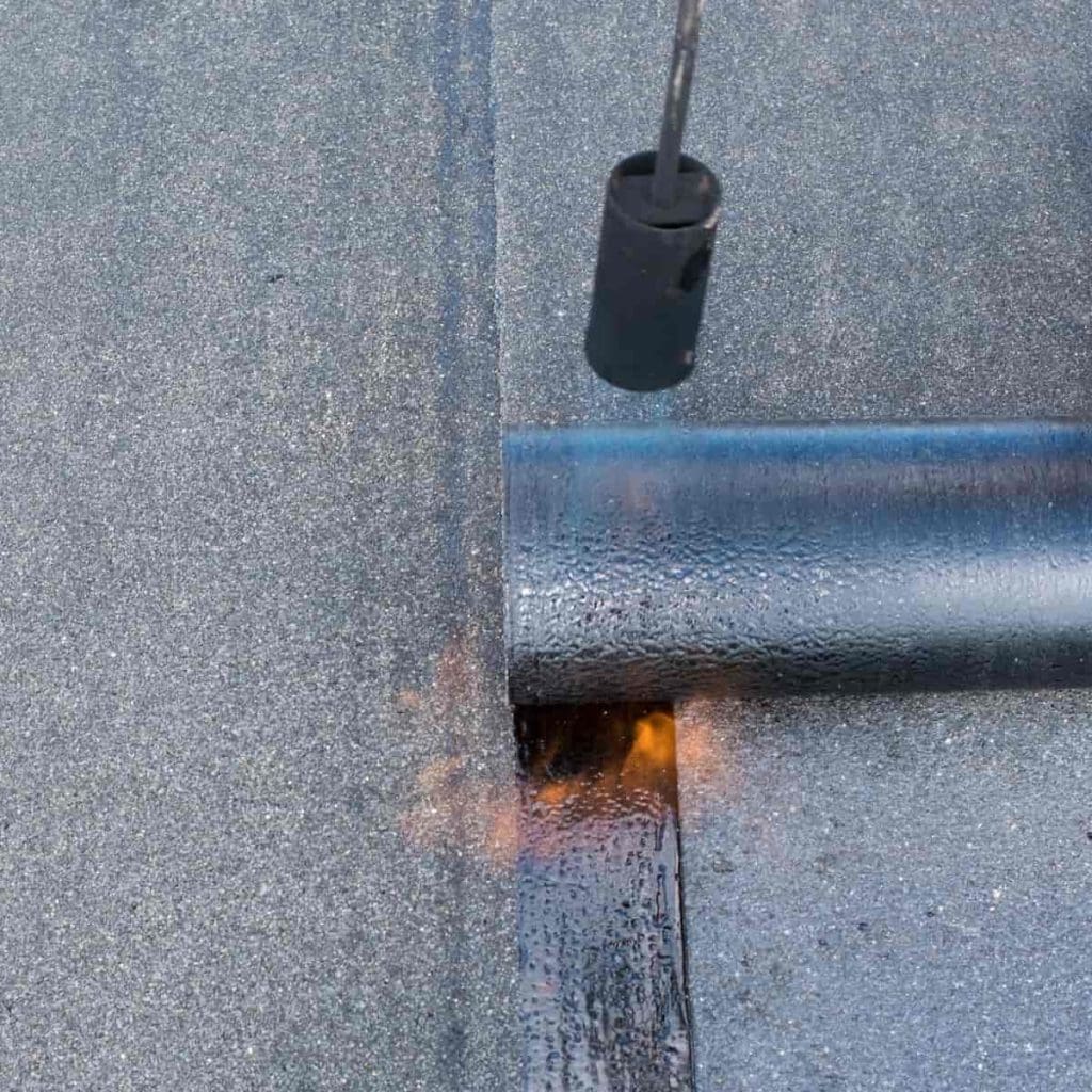 A roll of roofing felt being installed with a torch, which heats and bonds the material to the roof surface. A small flame is visible beneath the edge of the rolled material.