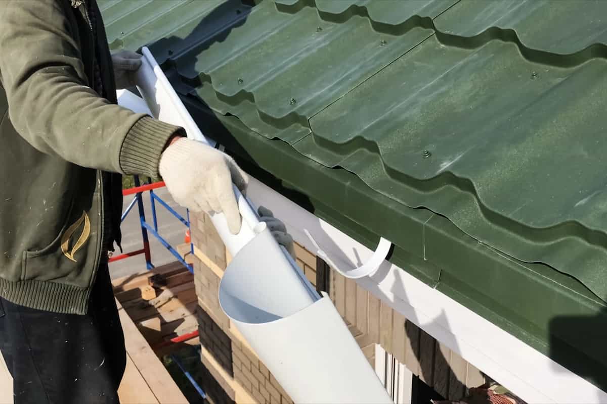 A person wearing gloves is installing a white rain gutter along the edge of a green metal roof on a house.