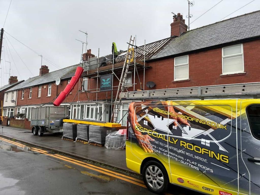 A row of brick houses under roof repair with scaffolding, a red debris chute, and a yellow Especially Roofing van parked on the street in front. A worker is visible on the roof.