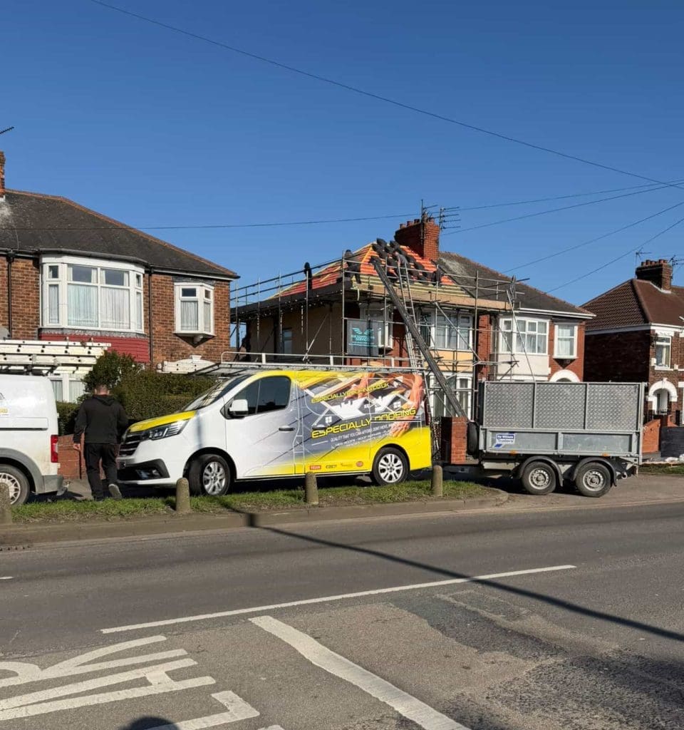 A house with scaffolding around the roof, workmen present, and a yellow van with a trailer parked outside. The van has roofing company graphics, and equipment is set up for roof repairs under a clear blue sky.