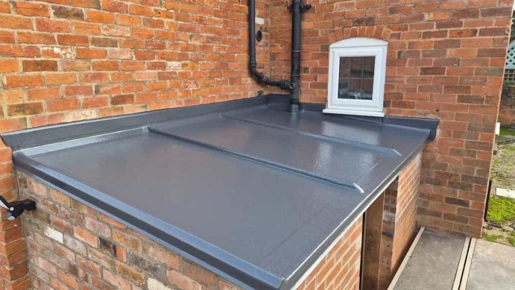 A flat roof extension with a smooth, dark grey waterproof surface is attached to a brick building, below a small white-framed window and surrounded by visible drainpipes.