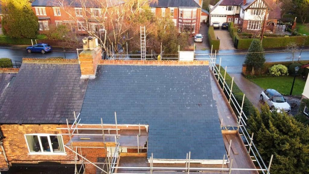 Aerial view of a house with scaffolding around a newly installed dark slate roof; older, weathered roof tiles are visible on the left side, with residential houses, trees, and a road in the background.