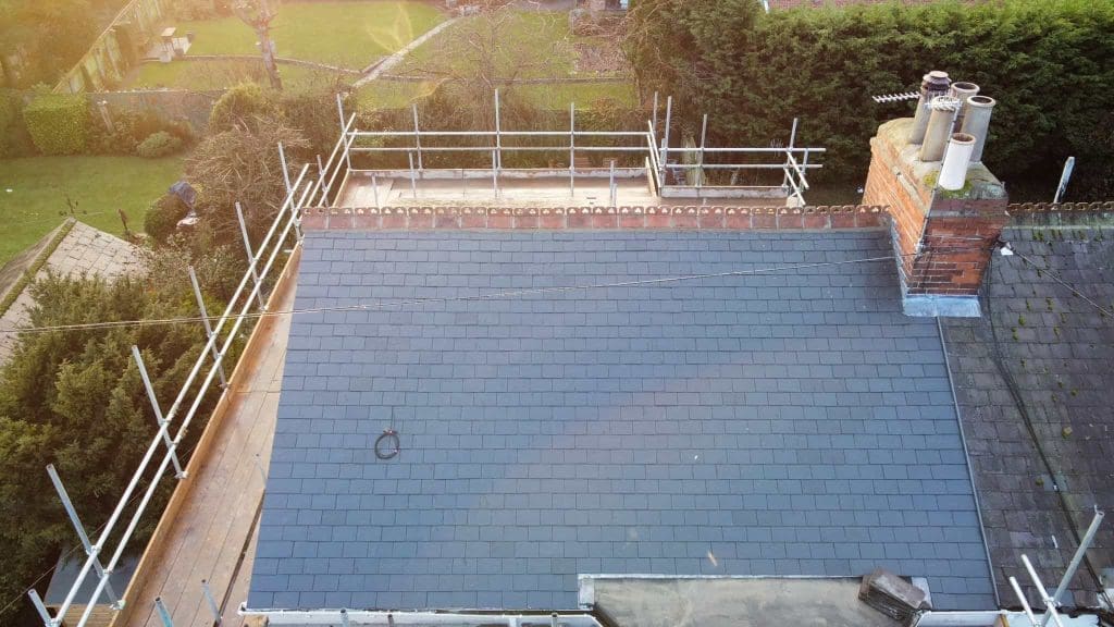 Aerial view of a newly tiled slate roof on a building, surrounded by scaffolding, with a brick chimney and garden areas visible in the background.