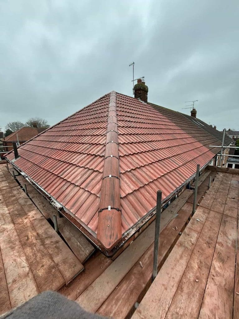 A newly tiled red roof on a house, viewed from above with scaffolding and wooden planks surrounding it, under a cloudy sky.