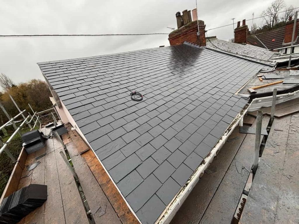 A newly installed slate roof on a house, viewed from above, with scaffolding surrounding the structure and a cloudy sky overhead. Some roofing materials and tools are visible on the scaffolding.