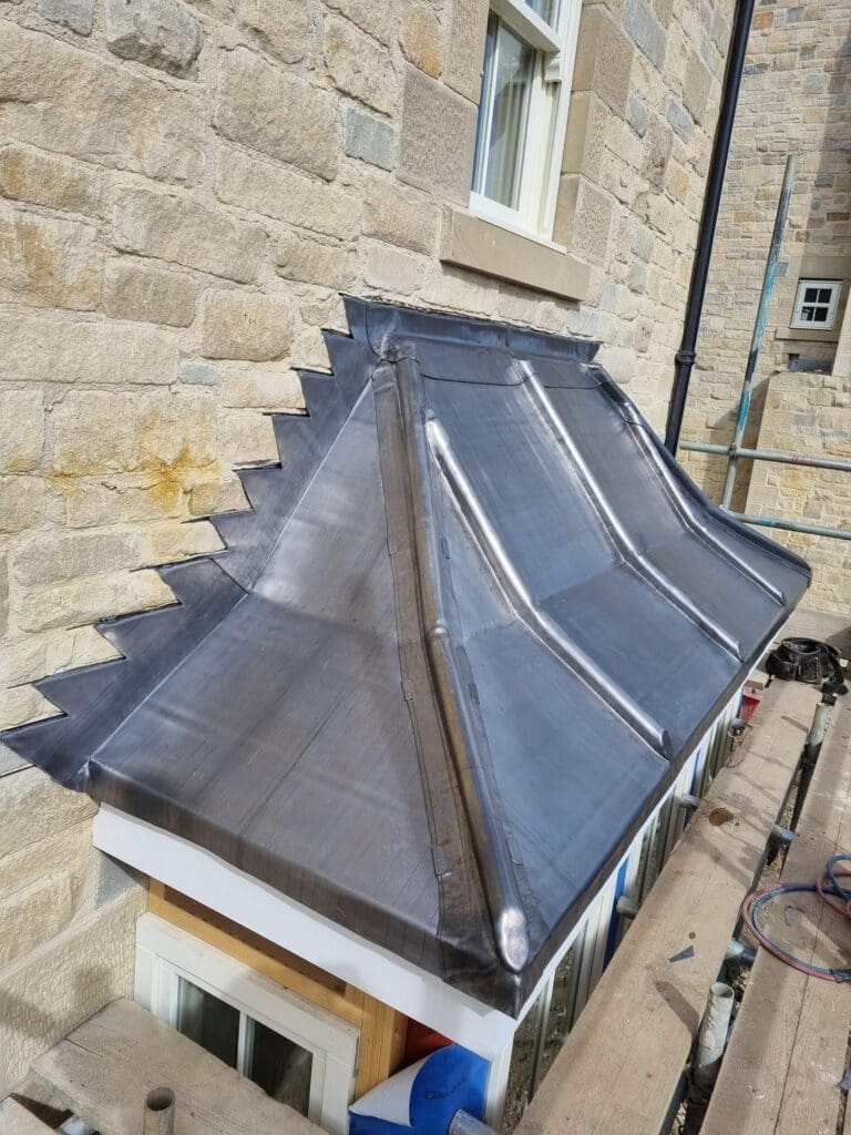 A newly installed lead roof with sharp, zigzagged edges covers a small extension beneath a stone wall with windows. Scaffolding and construction materials are visible nearby.