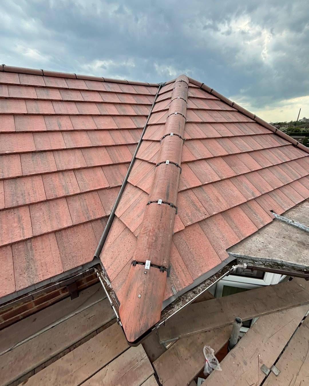 A close-up view of a red tiled roof with a ridge cap, taken from above. Scaffolding and cloudy sky are visible in the background.