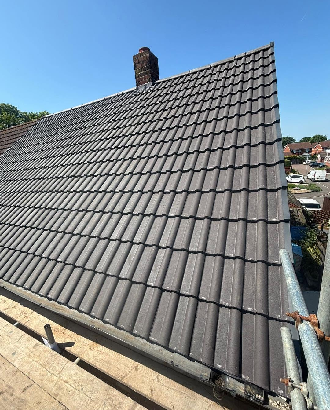 A close-up view of a gray tiled sloped roof with a chimney, taken from scaffolding on a sunny day; houses and trees are visible in the background.