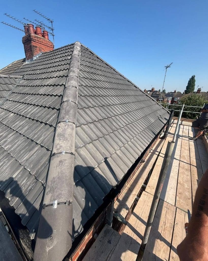A rooftop with grey tiles and a red brick chimney under a clear blue sky, seen from a scaffold platform. Rooftop antennas are visible in the background.