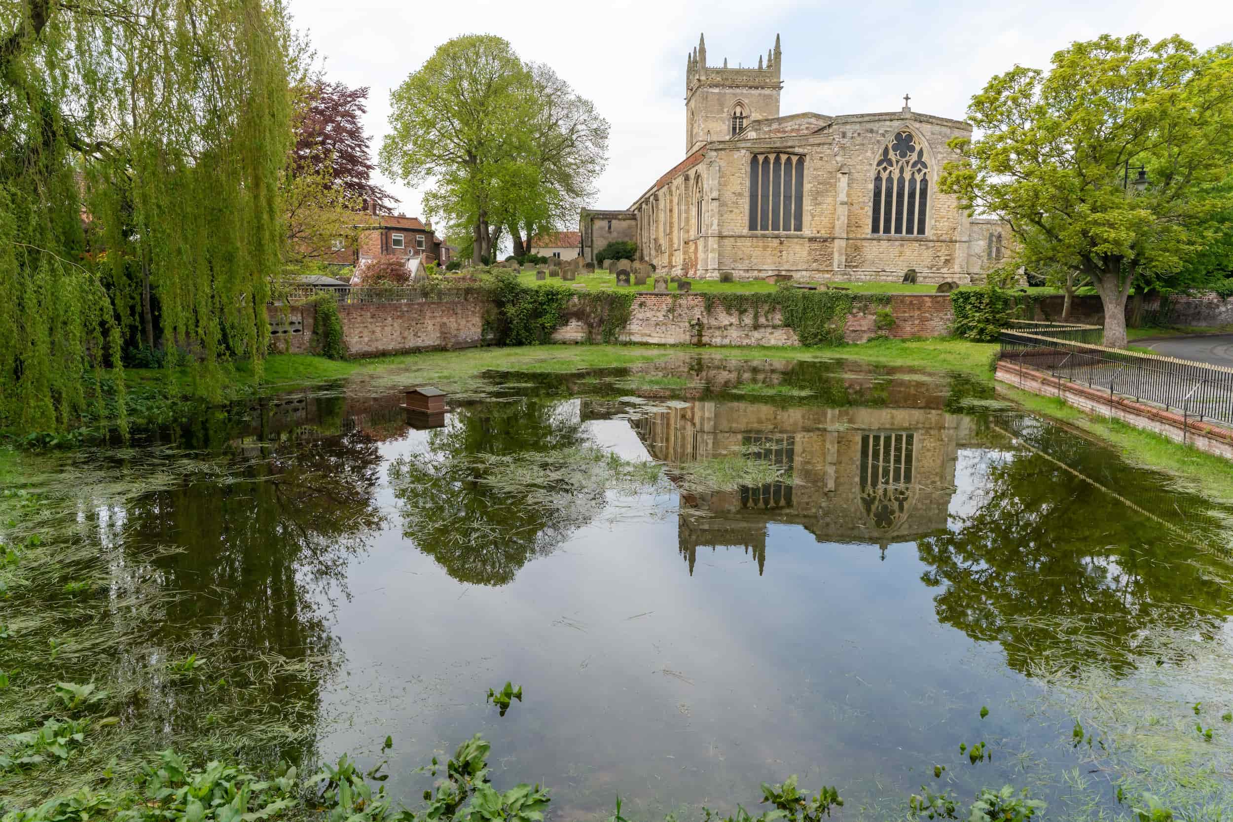A stone church with large windows sits beside a pond, surrounded by green trees. The church and trees are reflected clearly in the still water of the pond.