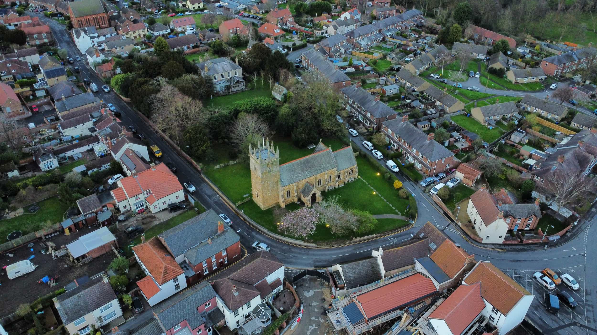 Aerial view of a village with a stone church at the center, surrounded by houses, gardens, roads, and parked cars. Trees and greenery fill the churchyard, with curved streets outlining the area.
