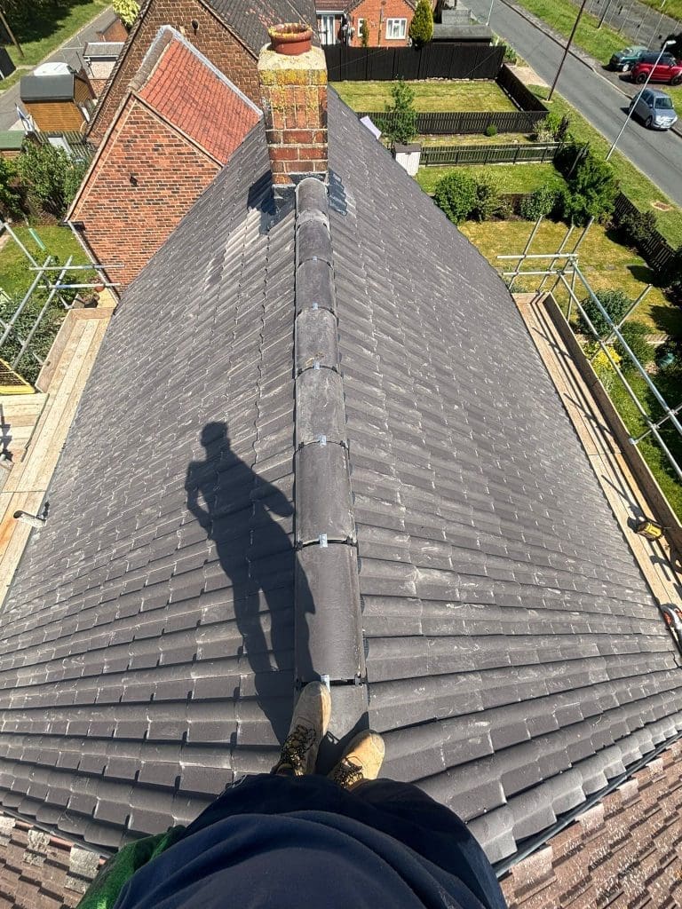 A person stands at the peak of a tiled roof, casting a long shadow. Scaffolding surrounds the house, and a chimney rises from the center of the roof. Green yards and nearby streets are visible below.
