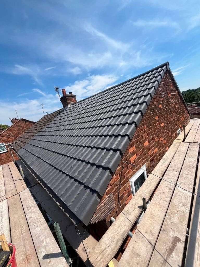 A brick house with new dark gray roof tiles, surrounded by scaffolding under a bright blue sky with scattered clouds.