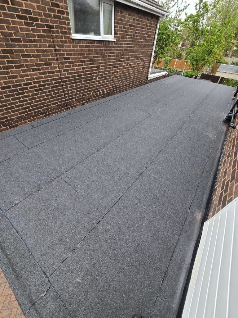 Flat roof with newly installed dark gray roofing felt on an extension, adjoining a brick house with a window. Trees and a street are visible in the background.