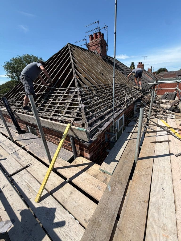 Two workers are repairing the wooden framework of a roof, with scaffolding and tools set up around the house under a clear blue sky.