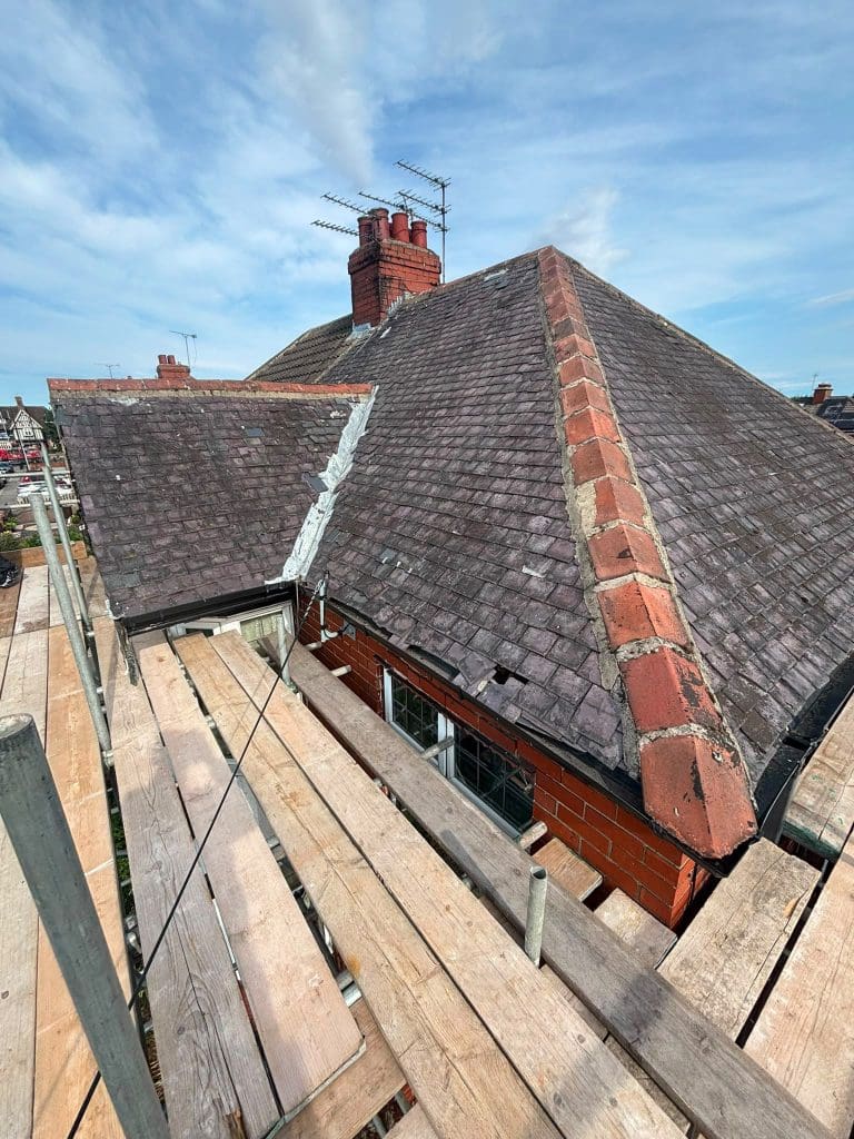 A rooftop with red and gray shingles, visible chimney, and scaffolding with wooden planks around the roof for repairs under a partly cloudy blue sky.
