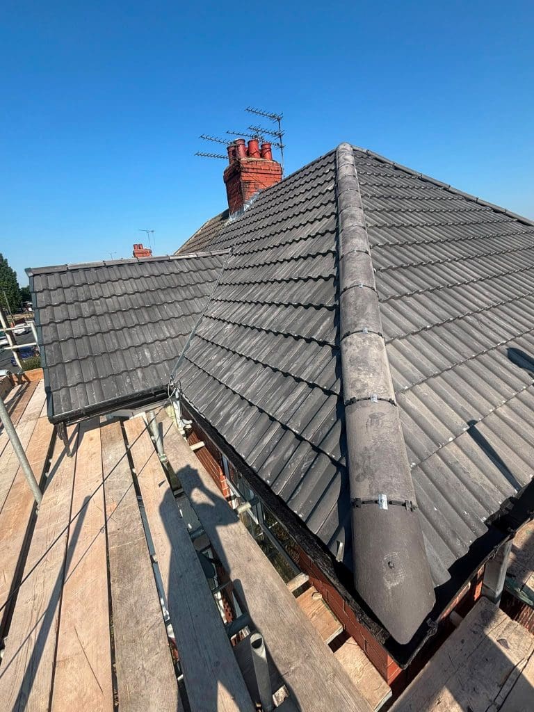 A view of a house roof with dark gray tiles and a brick chimney, taken from scaffolding on a sunny day with a clear blue sky.