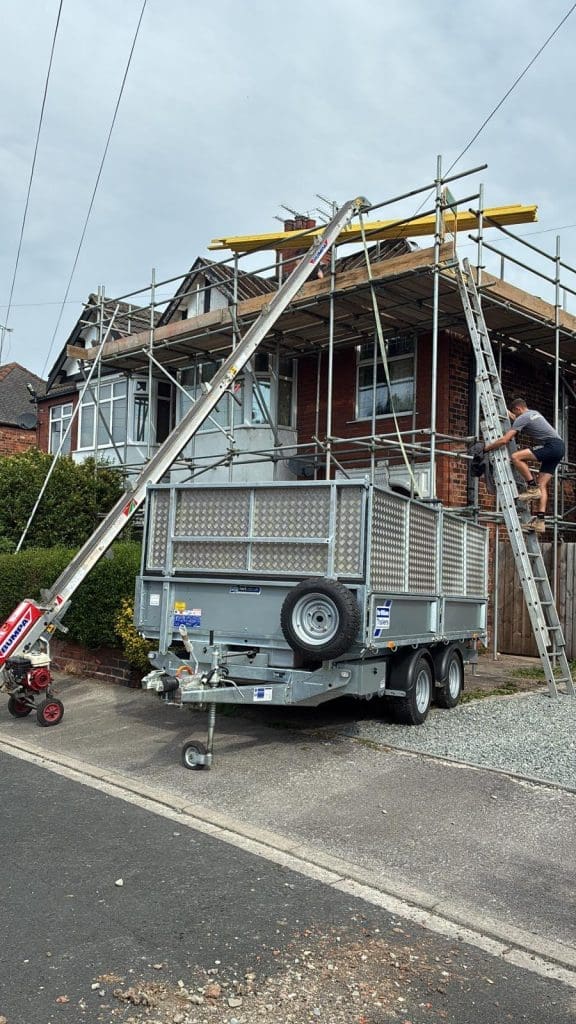A man climbs a ladder on scaffolding set up around a brick house under construction. A large metal trailer is parked in front, with equipment connecting to the scaffold above.