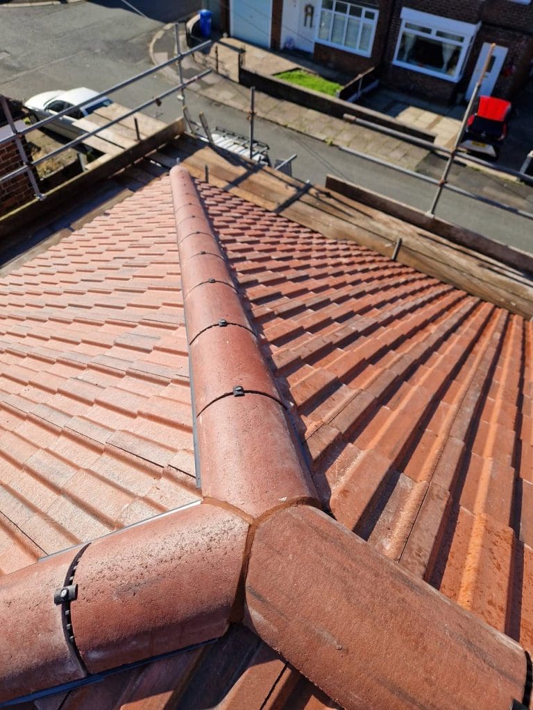 View from a rooftop shows red tiles and a ridge with clips, scaffolding along the roof edge, and a street below with parked cars, houses, and a ladder leaning against the scaffold.