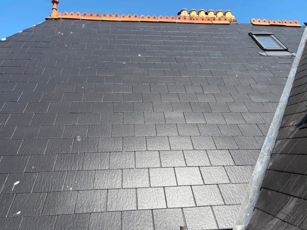 A black slate roof with clean rectangular tiles, an orange decorative ridge, multiple yellow chimney pots, and a small skylight on the right side against a clear blue sky.